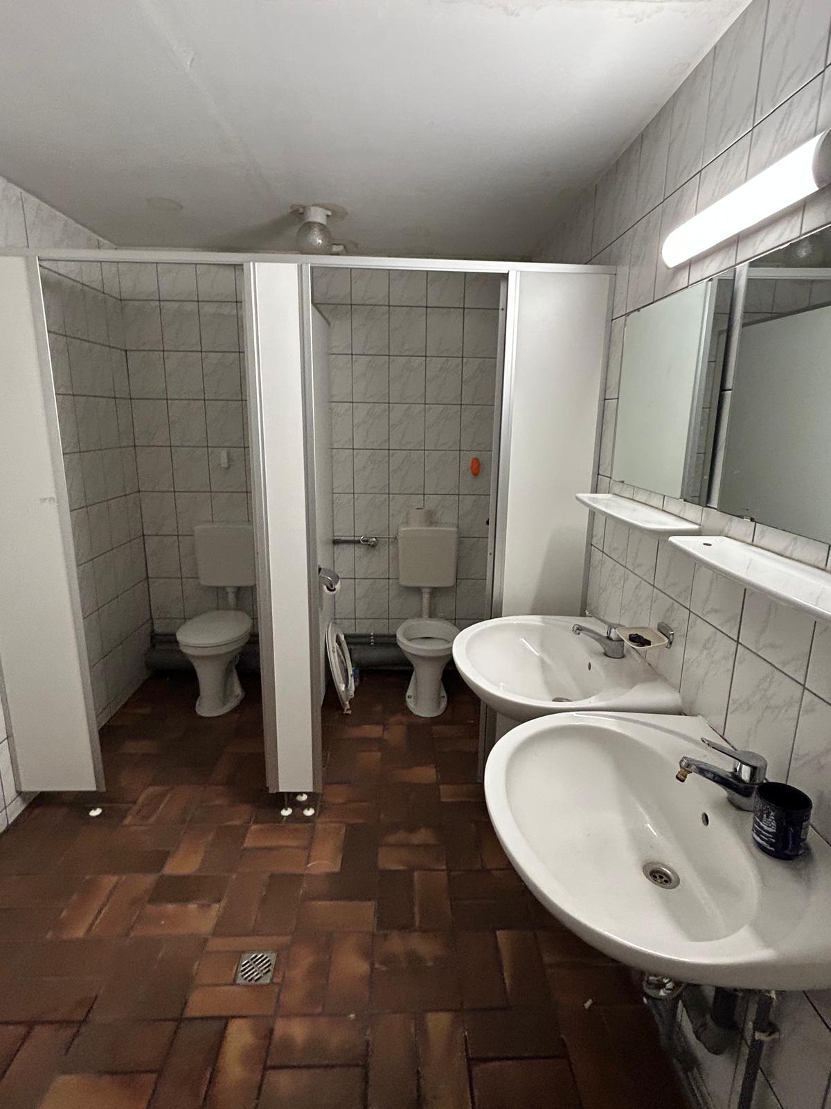 Alter gewerblicher Sanitärraum mit braunem Boden und älteren Wandfliesen