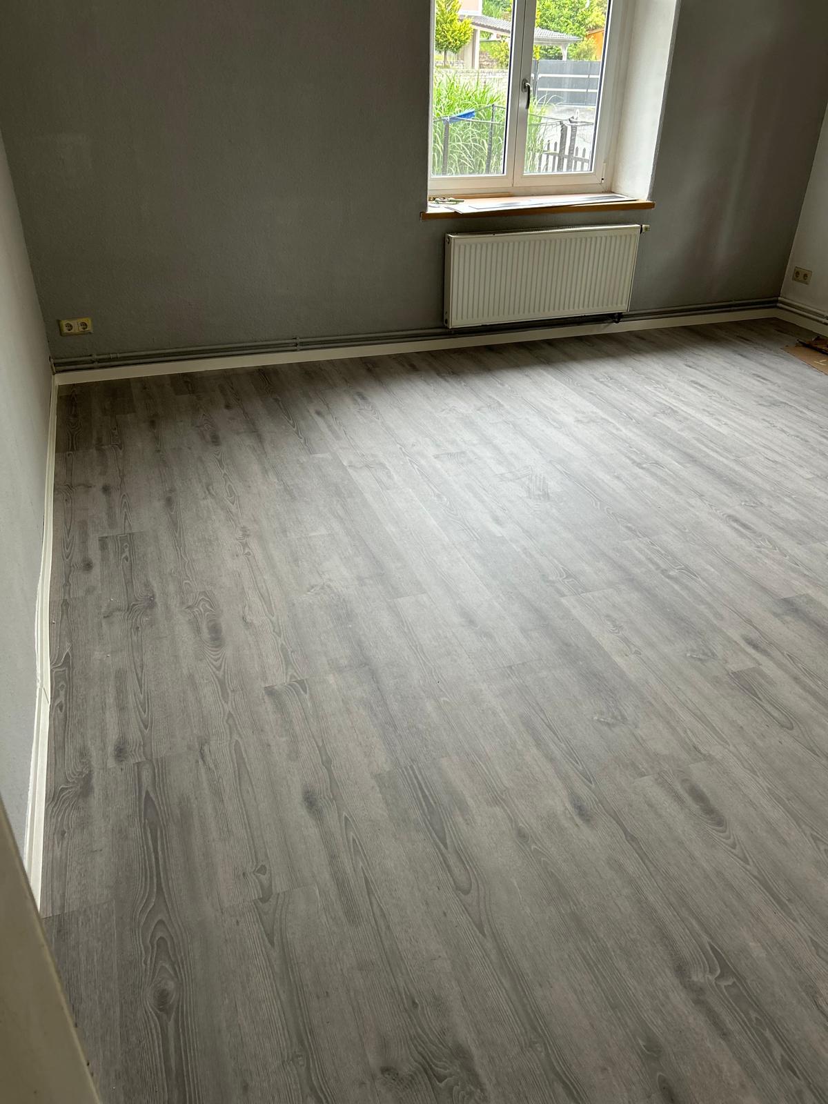 Wohnraum mit grauem Vinylboden und sauber geführten Anschlüssen an Wand und Heizungsrohre